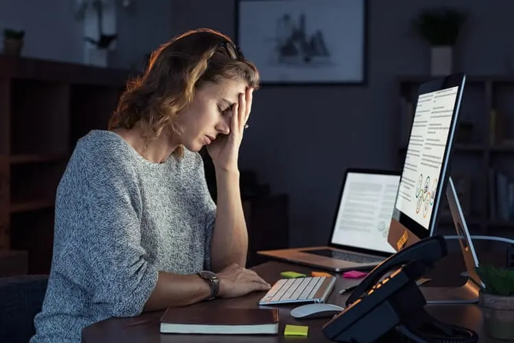 Mulher sentada em frente a mesa de trabalho, com computador, telefone, notebook, etc. Luzes apagadas, mão na cabeça, aparentemente cansada