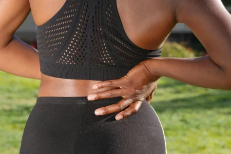 Uma pessoa de costas, vestindo roupas esportivas, segurando a região lombar com uma das mãos, indicando dor ou desconforto muscular.