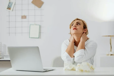 Mulher com dor no pescoço trabalhando no computador, sentindo desconforto e estresse.