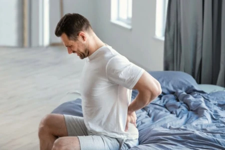 Detalhe de homem sentado na cama, com a mão nas costas, na região da lombar.