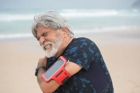 Homem na praia com expressão de dor intensa no ombro esquerdo, segurando o braço e o ombro com a mão direita.