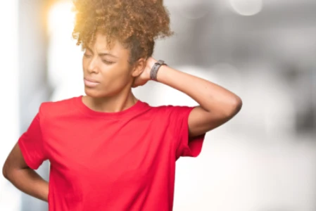 Mulher negra, com os cabelos presos, veste blusa vermelha e tem uma das mãos na parte de trás do pescoço. Ela está com expressão de dor.