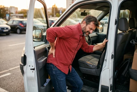 Homem de camisa vermelha e calça jeans azul escuro saindo do banco do motorista de um carro, segurando a região lombar com uma das mãos e exibindo uma expressão de dor e rigidez nas costas.