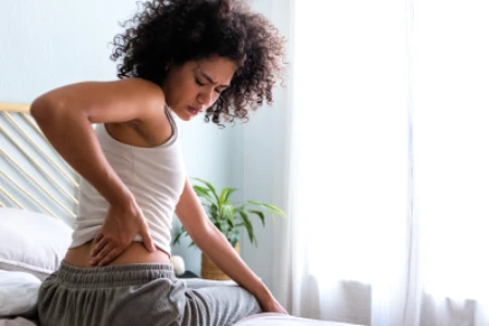 Mulher sentada na cama, com a mão direita tocando a coluna lombar, sentindo dores.