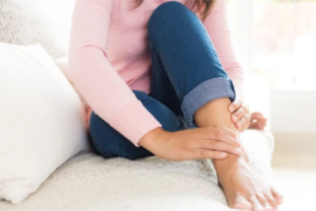 Detalhe das pernas de uma mulher, senta no sofá, com os pés também no sofá. Ela está massageando o tornozelo inchado com as mãos.