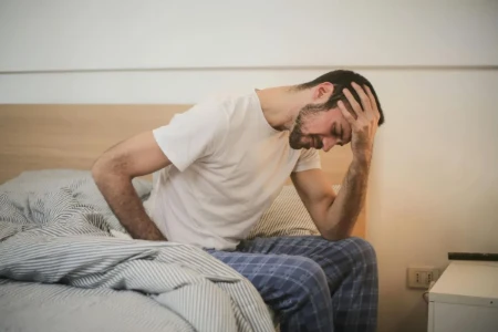 Homem sentado na beira da cama com expressão de dor intensa, com uma mão na cabeça e outra na região lombar, ilustrando um momento em que a dor no corpo se torna preocupante.