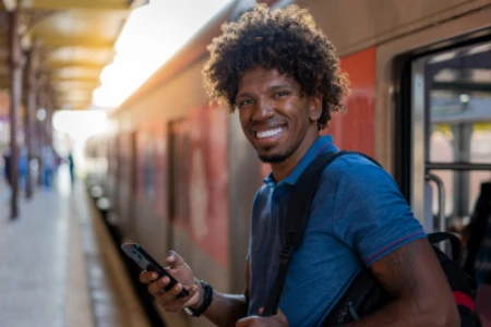 Rapaz sorrindo para a foto, segurando a alça da mochila com uma mão e um celular na outra, saindo do metrô.