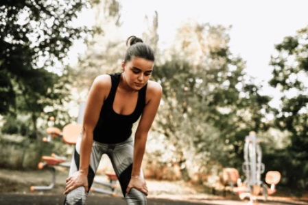 Mulher com roupas de academia, cabelo preso em coque, mãos apoiadas nos joelhos, em um parque bem arborizado.