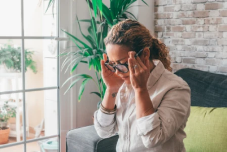 Mulher sentada em sofá está tocando suas têmporas, com expressão facial que revela dor.