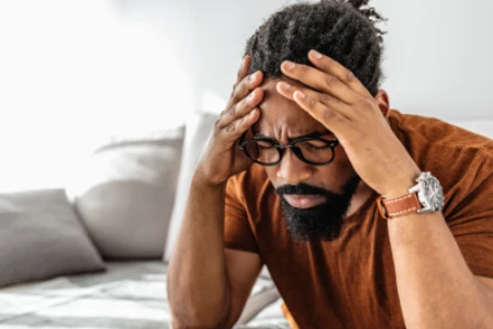 Homem sentado em sofá, com os cotovelos apoiados nos joelhos, as mãos segurando a cabeça que está abaixa - o rosto transparece dor.