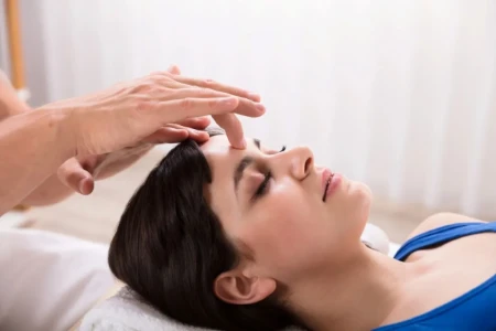Fotografia close-up de mãos de um terapeuta realizando uma massagem suave na testa e couro cabeludo de uma mulher deitada de olhos fechados.