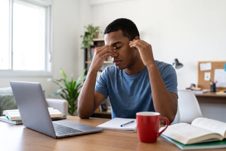 Homem de pele morena sentado em uma mesa de estudos com notebook, esfregando os olhos e a testa com expressão autêntica de cansaço visual e dor de cabeça. Uma caneca vermelha compõe a mesa.