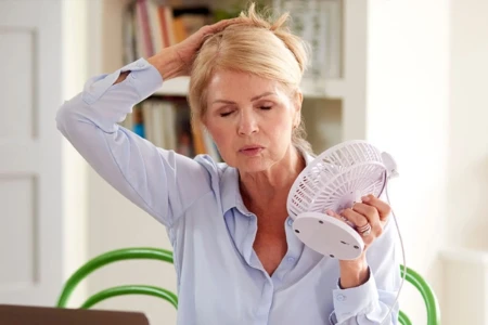 Mulher madura tocando a cabeça com uma mão, segurando um mini-ventilador com a outra, com olhos fechados e semblante de dor.