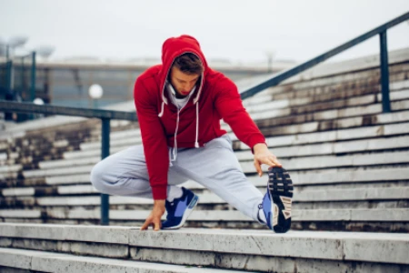 Homem de agasalho vermelho, alongando a perna e o pé esquerdo em arquibancada.