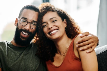 Um homem de barba e óculos e uma mulher de cabelos cacheados sorrindo e abraçados, transmitindo uma sensação de alívio, felicidade e conforto.
