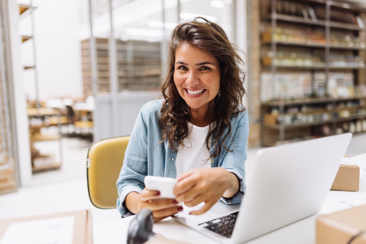Mulher sorridente sentada, segurando celular, em frente a um computador, em um depósito