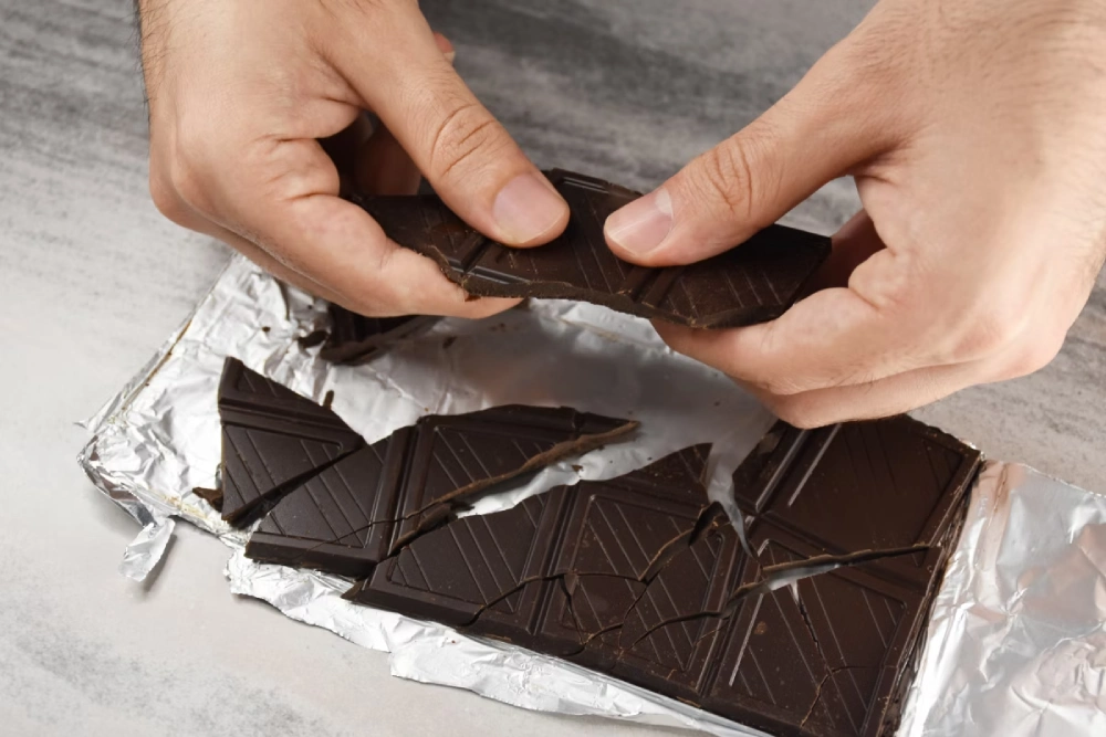 Detalhe de mãos quebrando uma barra de chocolate amargo em pedaços menores, sobre uma bancada e papel alumínio