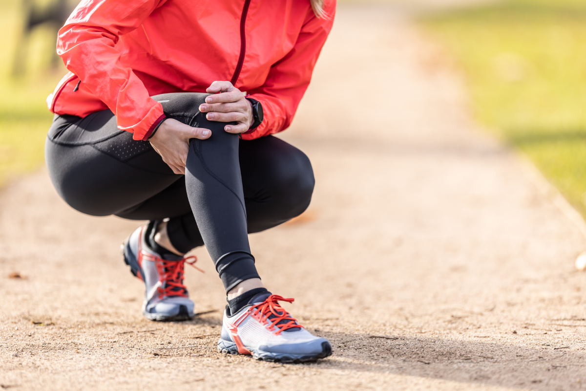 Close-up de uma mulher com jaqueta rosa e calça preta agachada em uma trilha de terra, segurando a canela e o joelho com as mãos, indicando dor muscular.