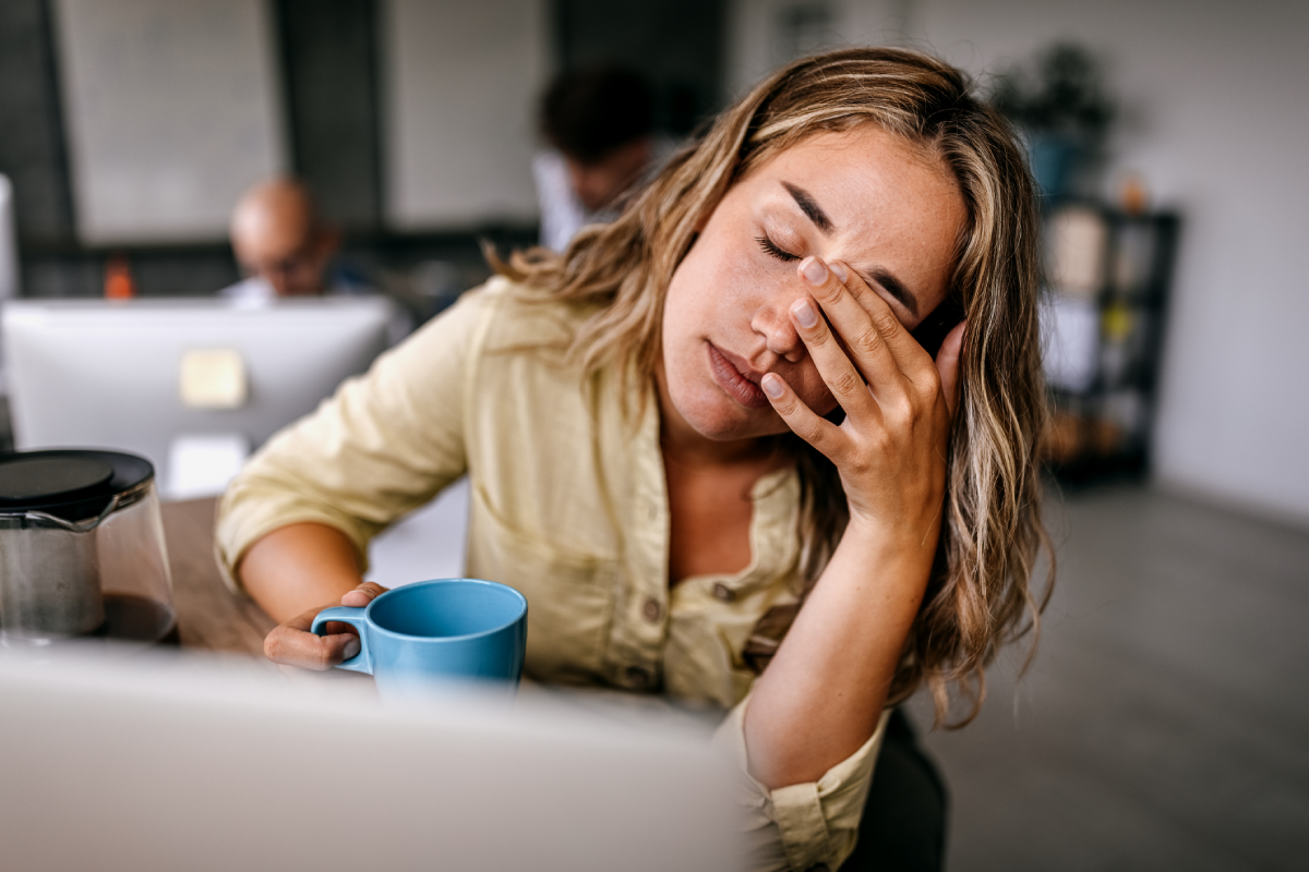Mulher sonolenta diante do computador. Ela segura uma xícara de café e leva uma das mãos à cabeça