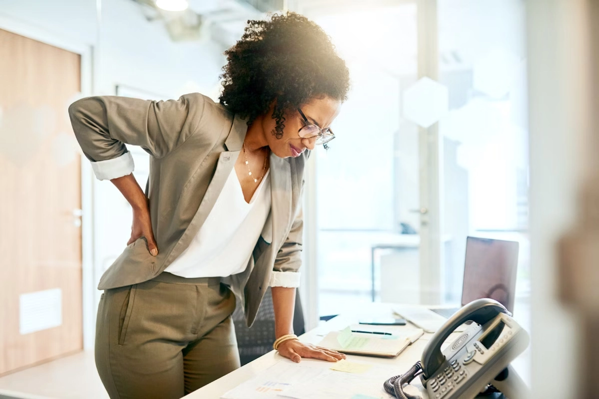Mulher no escritório apoiando uma mão na mesa, levemente curvada para a frente, com a outra mão apoiando a lombar