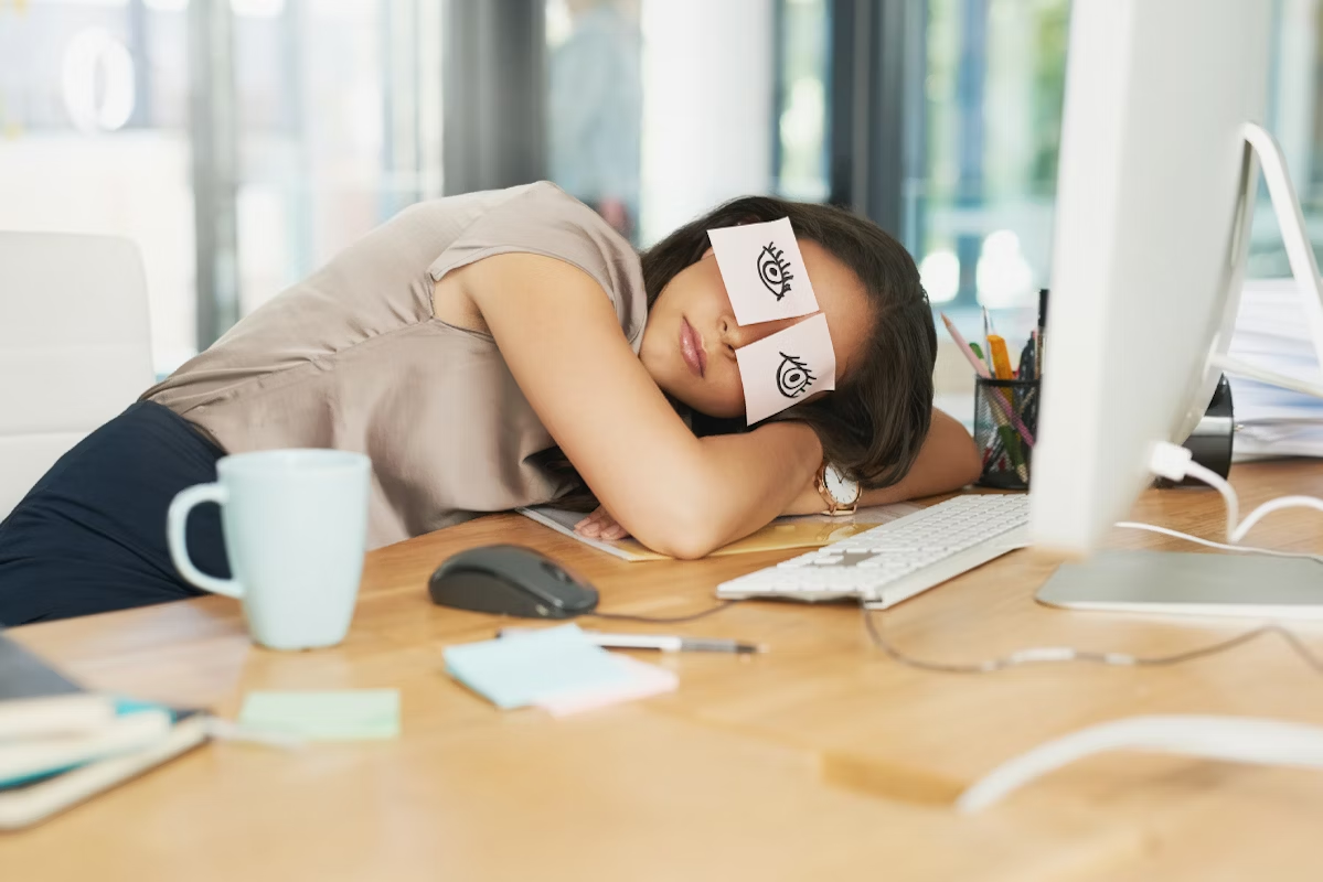 Mulher dormindo sobre a mesa de um escritório com posts com desenhos de olhos cobrindo os seus olhos.