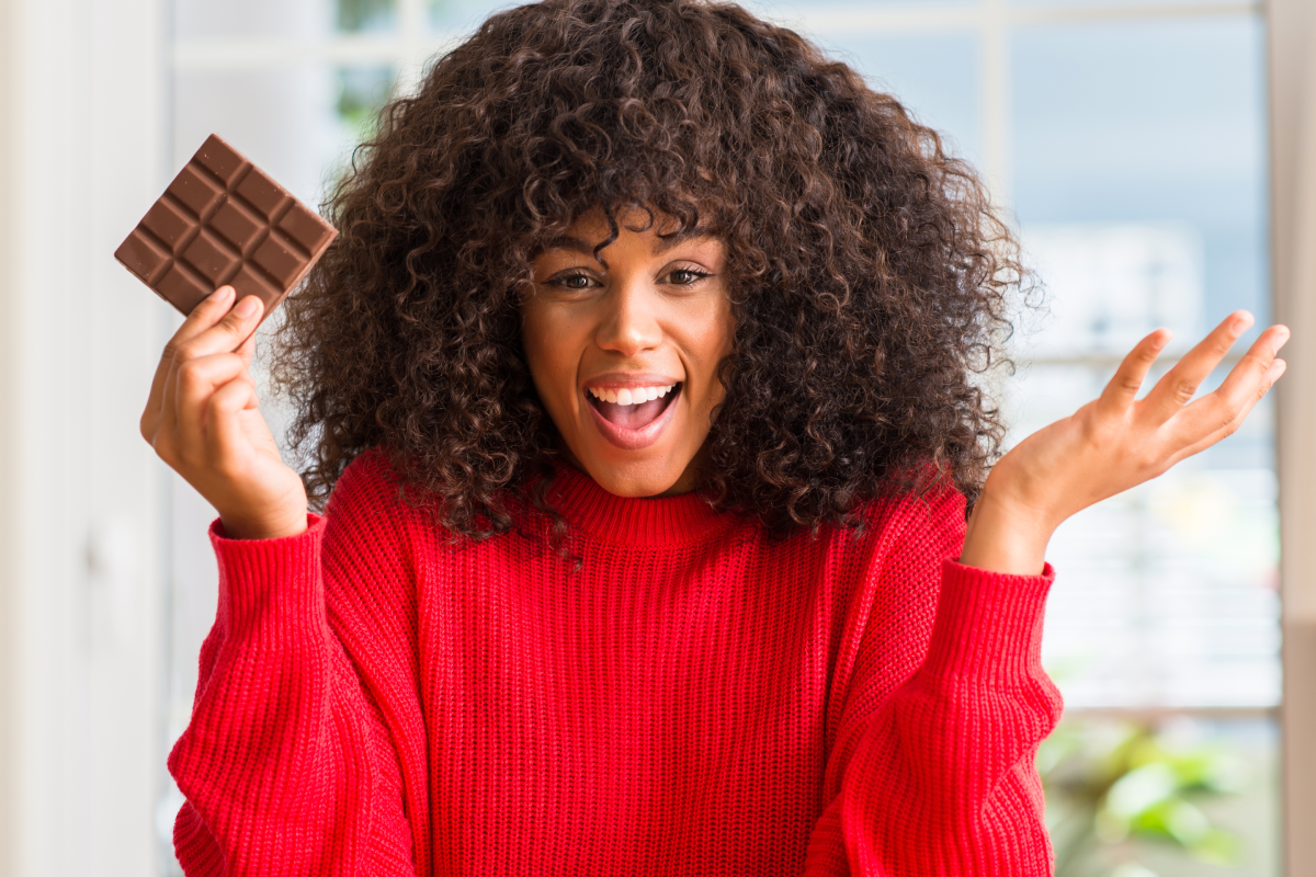 Mulher segurando chocolate, sorrindo, mas fazendo sinal de dúvida com as mãos