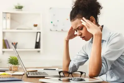 Jovem mulher no escritório, sentada em uma mesa com um laptop, segurando as têmporas com as duas mãos e com uma expressão de dor ou forte estresse/cansaço.
