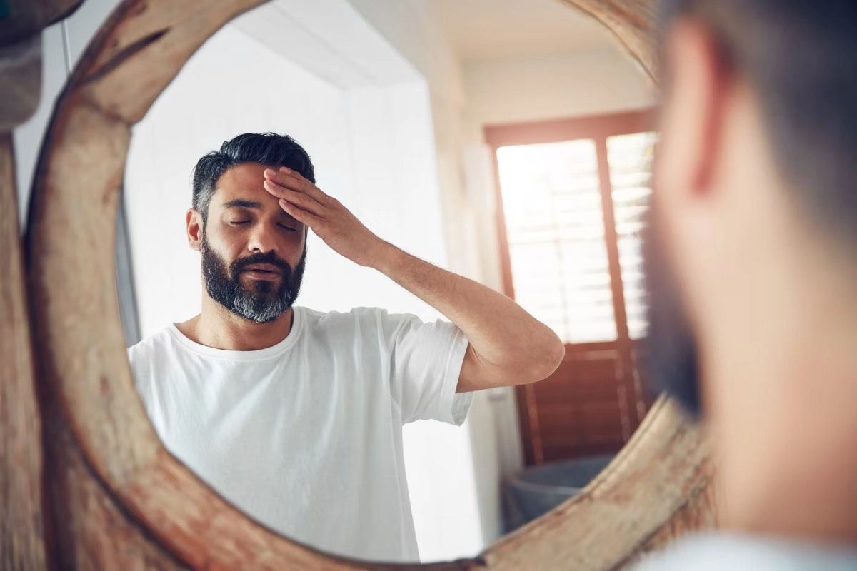 Homem de frente para o espelho, com olhos fechados e mão na testa, com feiçõs de quem sente dor