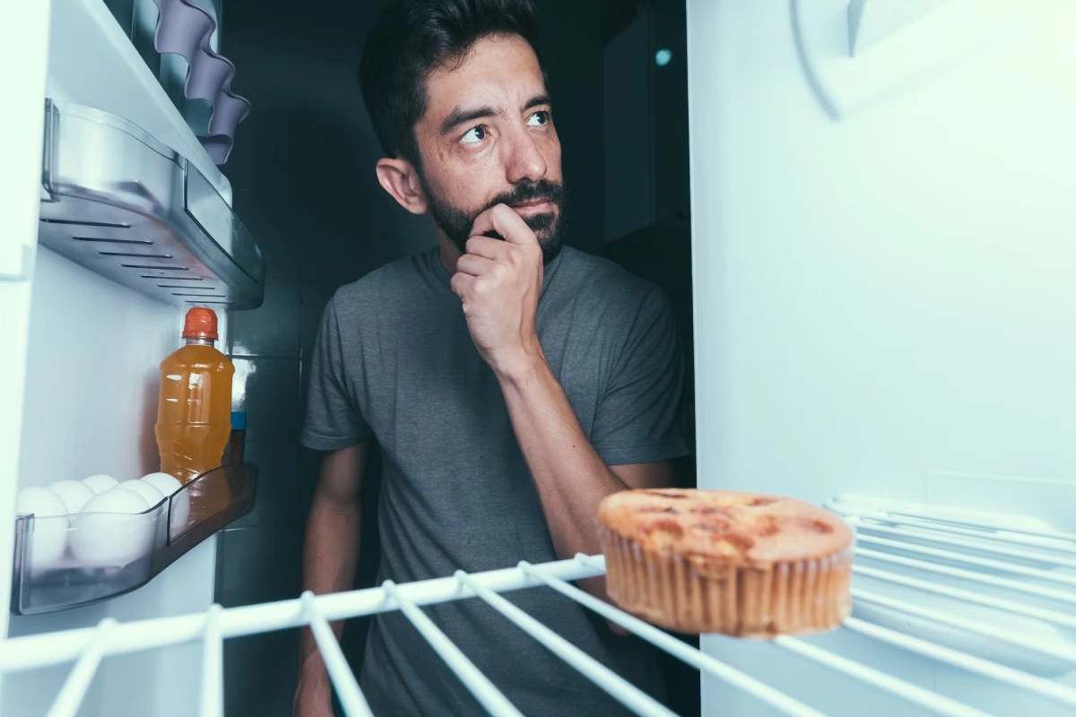 Homem com a geladeira aberta e a mão no queixo, refletindo se deve comer algo ou não