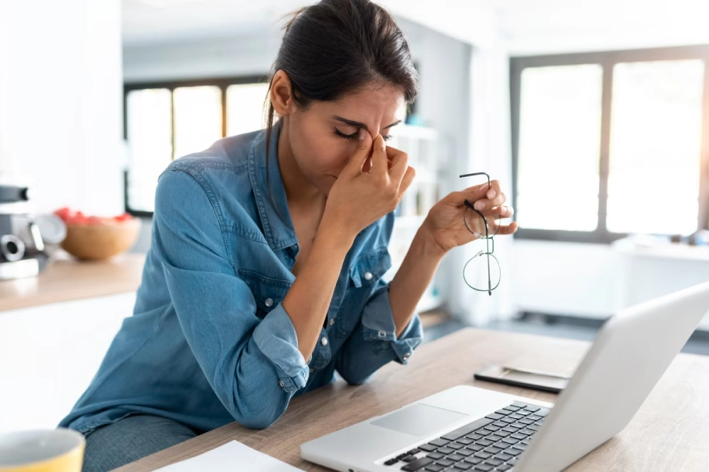 Mulher está sentada em frente a um notebook, segurando os óculos em uma mão e pressionando a região entre os olhos fechados com a outra.