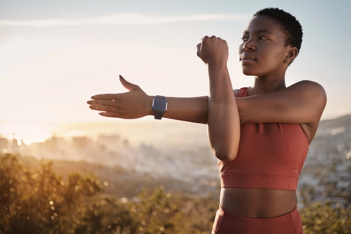 Mulher vestindo roupa de ginástica cor de ferrugem alongando o braço e o ombro direito em um cenário ao ar livre com o pôr do sol ao fundo.