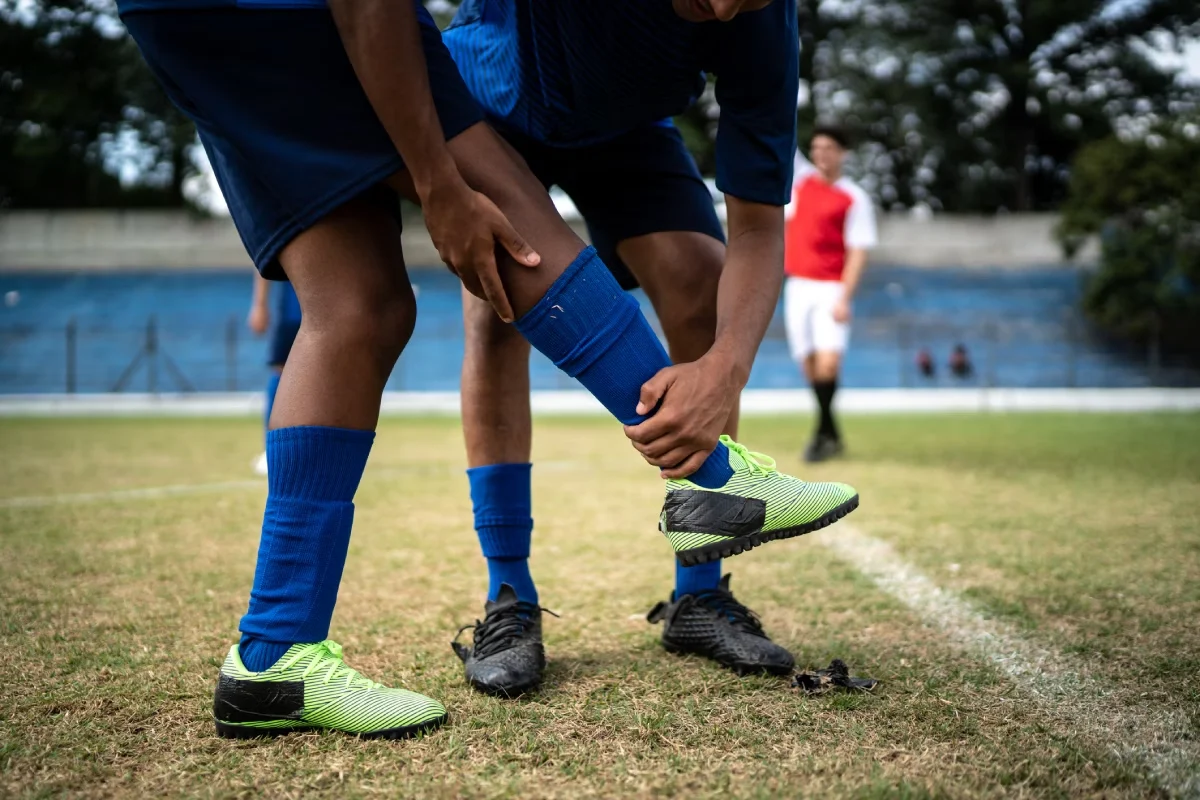 Jogador de futebol segurando e elevando a perna de um colega de time no campo para ajudar a alongar a panturrilha e aliviar a tensão muscular.