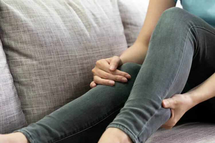 Mulher sentada com as pernas levemente dobradas em cima do sofá e com a mão na panturrilha.