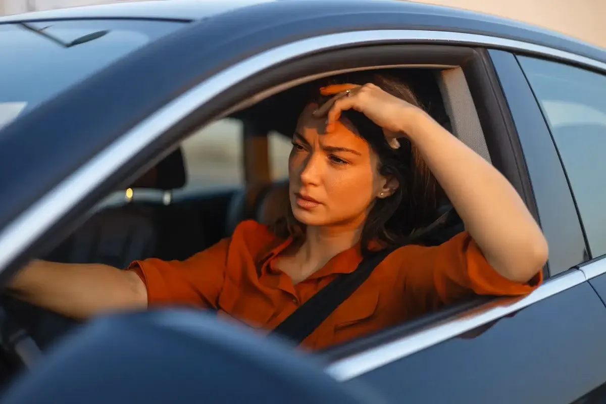 Mulher com a mão na cabeça sofrendo com exposição ao sol dentro do carro com a janela aberta
