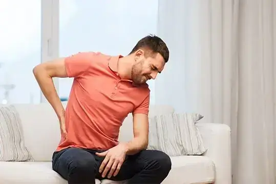 Homem sentado no sofá com a mãp na lombar sentindo dores