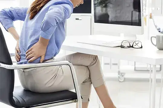 Mulher sentada na frente do computador com as mãos na lombar sentido dor