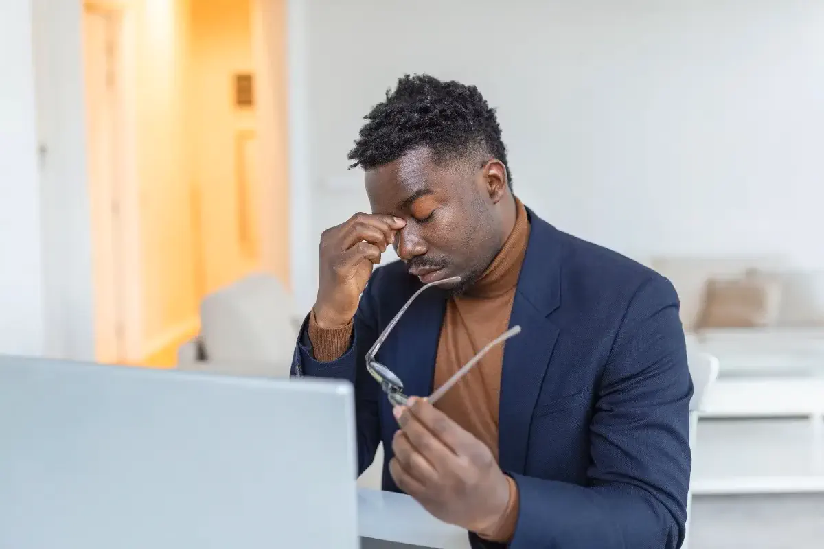 Homem em ambiente de trabalho, sentado na frente do computador massageando acima do nariz e com óculos na outra mão