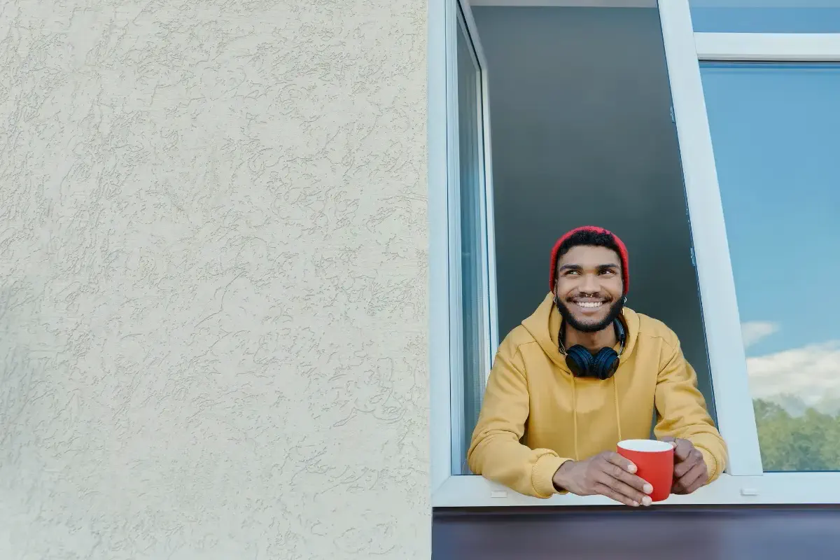 Homem na janela com uma xícara na mão, sorrindo ao respirar ar fresco